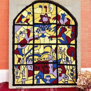 Op de afbeelding: Een kleurrijk wandkleed van naaldvilt dat een glas-in-loodraam voorstelt met een Davidster, Hebreeuwse tekst en andere religieuze symbolen.