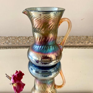 May include: A iridescent glass pitcher with a swirl design and a clear amber handle. The pitcher is sitting on a mirrored surface with a single pink rose.