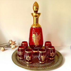 以下が含まれることがあります： 金色のアクセントと宗教的なデザインが施された赤いガラスのデカンタと、それに合わせた6つのショットグラスが、真鍮のトレイに乗っています。