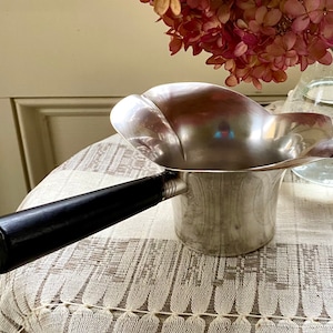 May include: A silver creamer with a black wooden handle. The creamer has a unique, curved shape and a shiny finish.