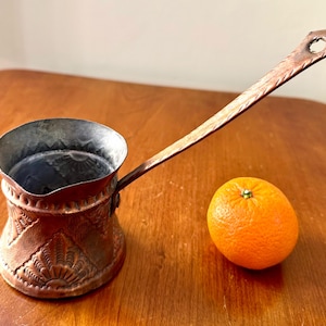 May include: A copper Turkish coffee pot with an ornate design and a long handle. The pot is sitting on a wooden surface next to a whole orange.