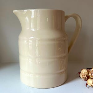 May include: A cream-colored ceramic pitcher with a handle and spout. The pitcher has horizontal ridges around the body. Dried roses are in the lower right corner. The pitcher is on a white surface.