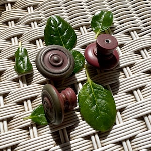 Puede incluir: Vista cenital de cuatro tiradores decorativos para armarios en tonos burdeos y grises, dispuestos con hojas verdes sobre una superficie tejida blanca. Los tiradores tienen un diseño circular con un centro en relieve y son de material sólido.