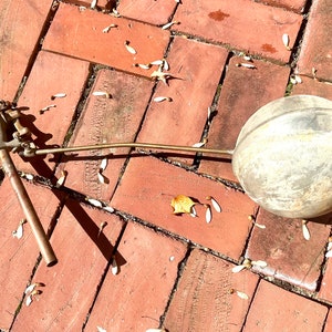 Puede incluir: Una válvula de flotador de tanque de agua de latón vintage con una bola de flotador redonda de color plateado. La válvula está sobre un patio de adoquines con pétalos de flores blancas esparcidos alrededor.