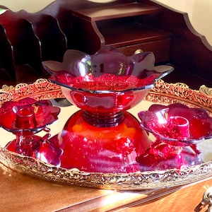 May include: A set of three ruby red glass bowls on a gold mirrored tray. The bowls are in a flower shape and have a footed base. The tray has a decorative gold rim.