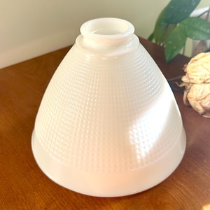 May include: A white, cone-shaped glass lampshade with a textured, dotted pattern. The lampshade has a wide base and a narrow opening at the top. It is sitting on a wooden surface.