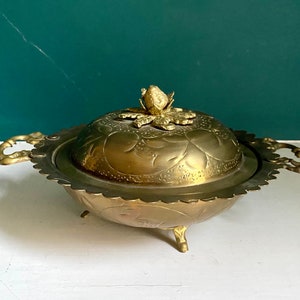 Puede incluir: Un plato de servir de latón dorado con tapa. La tapa tiene un diseño floral y un pomo decorativo en el centro. El plato tiene un borde festoneado y tres patas.