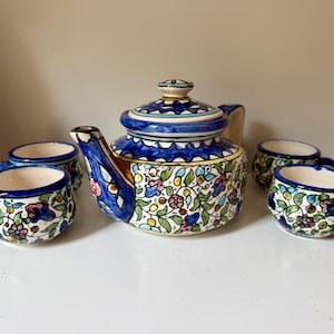 May include: A ceramic tea set featuring a teapot and four teacups. The set is decorated with a floral pattern in blue, green, yellow, and pink. The teapot has a blue lid and handle. The teacups have blue rims and handles.