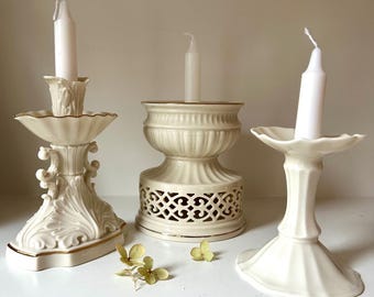 May include: Three ivory-colored ceramic candle holders with white candles. The candle holders have ornate designs, including floral and geometric patterns. Gold trim accents the edges. Dried flowers are scattered in front of the candle holders.