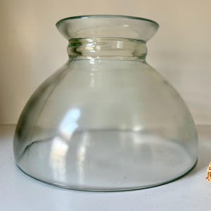 Op de afbeelding: Een heldere glazen kom met een afgeronde basis en een uitlopende rand. De kom heeft een breed, open ontwerp en is geschikt om te serveren of tentoon te stellen. Het glas is glad en reflecteert licht. Gedroogde bloemen zijn zichtbaar op de achtergrond.