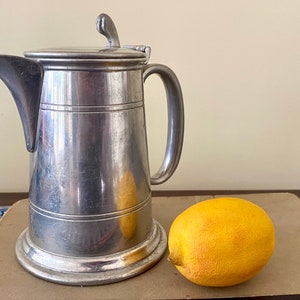 Op de afbeelding: Een zilveren tinnen kan met een deksel en een handvat. De kan staat op een bruin oppervlak naast een gele citroen.