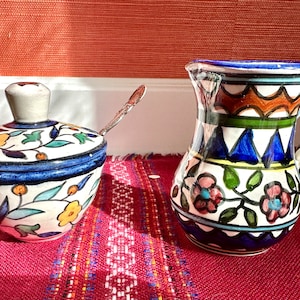 May include: A set of two hand-painted ceramic kitchenware pieces. A white ceramic sugar bowl with a lid and floral pattern in blue, green, and yellow. A white ceramic pitcher with a handle and floral pattern in blue, green, pink, and orange.