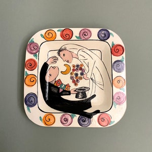 May include: A white ceramic square plate with a painted design of a bride and groom. The bride is wearing a white dress and the groom is wearing a black suit. The plate is decorated with a border of colorful flowers.