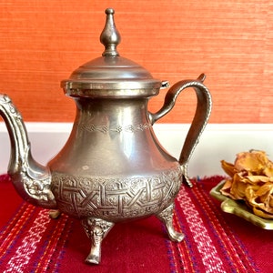 May include: A silver teapot with an ornate design and a bird-shaped spout. The teapot has three legs and a lid with a finial. The teapot is sitting on a red and white striped rug.