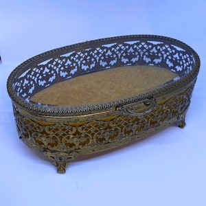 May include: An oval-shaped, gold-toned trinket box with a glass top and intricate floral designs. The box has four feet and a decorative handle.