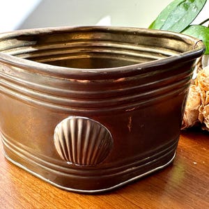 Może przedstawiać: Wazon w stylu vintage w kolorze brązu z muszelkowym akcentem. Donica ma prostokątny kształt z zaokrąglonymi rogami i poziomymi żłobieniami. Muszla jest wyśrodkowana z przodu. Donica stoi na drewnianej powierzchni.