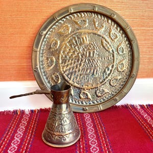 May include: An antique brass coffee pot with a long handle and a decorative, engraved design. A large, round brass tray with an embossed pattern is behind the pot. The items are on a red and white striped textile.