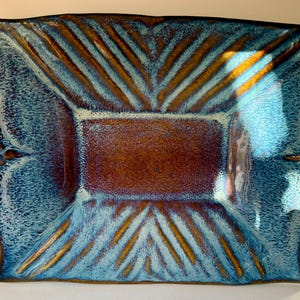May include: A rectangular ceramic serving platter featuring a deep brown center and vibrant blue glazed edges. The surface displays a textured pattern of raised diagonal lines radiating from the central rectangle toward the outer rim.