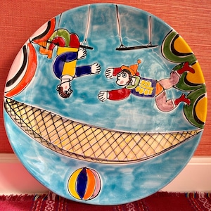 Può includere: Un piatto in ceramica blu con un'illustrazione colorata di due figure su altalene, una rete e una palla. Le figure sono vestite con costumi colorati e hanno tratti esagerati.