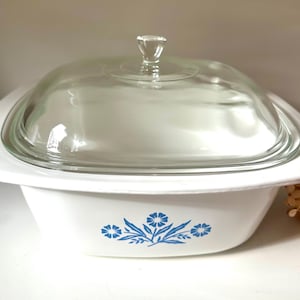 Puede incluir: Fuente para horno blanca con tapa de cristal transparente. El plato tiene forma cuadrada con bordes redondeados y un diseño floral azul. La tapa tiene un tirador de pomo transparente. Un artículo de cocina vintage.