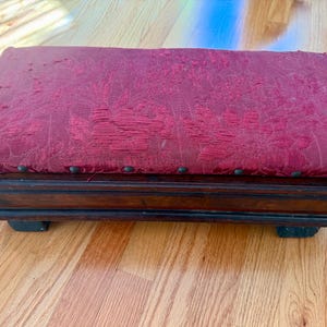 May include: An antique footstool with a rectangular shape. The top is upholstered in a faded red fabric with a floral pattern. The wooden frame is dark brown with decorative details and small, dark studs along the edge.