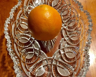 May include: A clear glass bowl with a decorative cut-glass pattern. An orange sits in the center of the bowl.