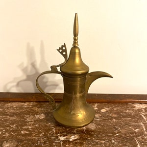 May include: A gold-colored brass teapot with a spout and a handle. The teapot has a decorative design on the body and a pointed lid.