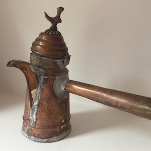 Puede incluir: Una cafetera antigua de cobre con un pico largo y un adorno decorativo en forma de pájaro. La cafetera tiene un cuerpo cilíndrico y un asa. El cobre tiene una pátina, que muestra signos de edad y uso. La cafetera probablemente se utiliza para preparar café.