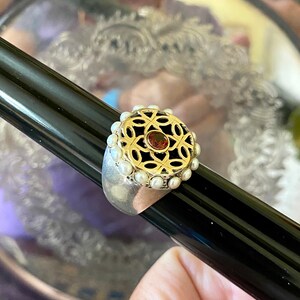 May include: A silver ring with a gold filigree design and a red gemstone in the center. The ring is surrounded by a row of white pearls.