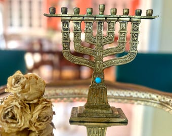 May include: A gold-coloured menorah with nine candle holders. The menorah features detailed designs and a turquoise stone at the base. Dried flowers are in the foreground. The menorah is a symbol of the Jewish faith.