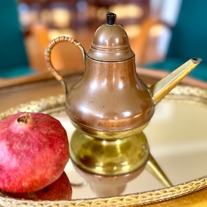 May include: A copper teapot with a brass base and a woven handle sits on a gold tray with a mirrored surface. A red pomegranate is also on the tray.