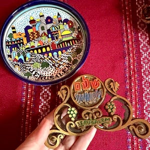 Könnte beinhalten: Ein Serviettenhalter aus Messing mit einem Jerusalem-Motiv. Der Halter verfügt über ein Weinrebenmotiv und die Worte "Shalom" und "Jerusalem" in Hebräisch und Englisch.