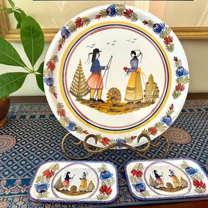 May include: A set of decorative ceramic serving pieces featuring a hand-painted design. The set includes a large round tray and two rectangular trays, all with a central image of figures in traditional attire. The rims are adorned with floral and geometric patterns.