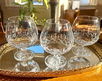 May include: Three clear glass brandy snifters with a cut crystal design sit on a gold mirrored tray.