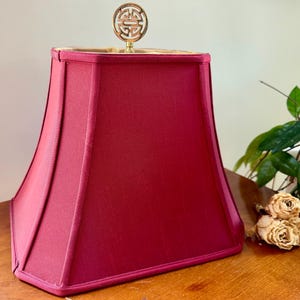 May include: A rectangular, deep red fabric lampshade with a structured, tapered silhouette and piped edges. The top center features a circular, metallic gold finial with an intricate, openwork geometric design, providing a decorative accent.
