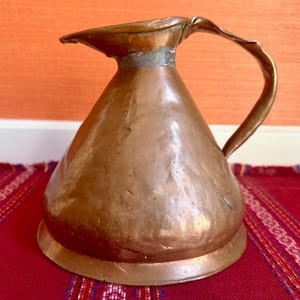 May include: A hammered copper pitcher with a wide, conical base, a narrow neck, a prominent pouring spout, and a curved handle. The metal surface displays a warm, metallic brown tone with visible indentations from the hand-hammering process.