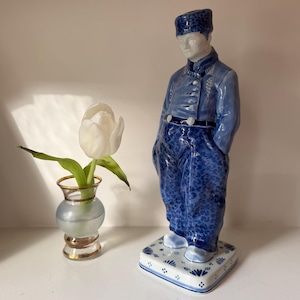 May include: A ceramic figurine of a man in a blue uniform with white accents. The figurine stands on a square base with a floral pattern. A small vase with a white tulip is to the left of the figurine.