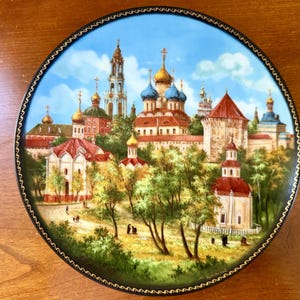 May include: A round decorative plate featuring a detailed painting of a cityscape with churches and buildings. The plate has a black border with a gold trim. The sky is blue, and the buildings have gold and blue domes.