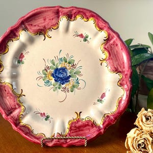 Könnte beinhalten: Dekorative Keramikplatte mit gewelltem Rand und handgemaltem Blumendesign. Die Platte hat eine cremefarbene Mitte mit einer blauen Rose und grünen Blättern. Der Rand ist in einem tiefen Rosa mit goldenen Akzenten bemalt. Ein Vintage-Stück.