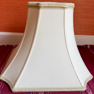 May include: A cream-colored bell-shaped lampshade with a square base and rounded corners. The shade features a smooth fabric surface with vertical piping along the edges and a decorative braided trim along the top and bottom borders.