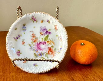 May include: A white porcelain plate with a gold rim and floral design. The plate is on a gold metal stand. A bright orange orange is sitting on a wooden surface.