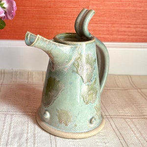Puede incluir: Una jarra de cerámica verde claro con un asa única y un esmalte moteado. La jarra tiene una base redonda y un pico.