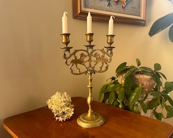 Può includere: Un candelabro in ottone dorato con tre portacandele e un design decorativo con animali stilizzati. Il candelabro è su un tavolo di legno con un arrangiamento di fiori bianchi.