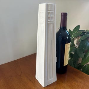 May include: A white ceramic vase with a geometric design. The vase is tall and slender, with a square base and a rectangular opening at the top. The vase is sitting on a wooden table.