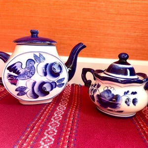 May include: A white and blue ceramic teapot and sugar bowl set. The teapot features a blue bird and floral design. The sugar bowl has a matching floral pattern. Both pieces have blue accents and handles. The set is on a red patterned tablecloth.