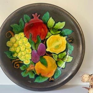 May include: A ceramic plate with a dark brown background and a colorful fruit design. The design features a red pomegranate, yellow lemons, green grapes, and purple plums.