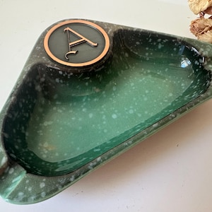 May include: A green ceramic ashtray with a speckled finish. The ashtray has a raised, circular section with a copper-colored letter "A" in the center.