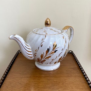 May include: White teapot with gold leaf design. The teapot has a round body and a lid with a knob on top. The teapot is sitting on a wooden surface.