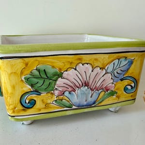 May include: Rectangular ceramic planter with a yellow background and a floral design. The flower is pink and blue with green leaves. The planter has a green rim and small white feet. Blue handles are on the sides.