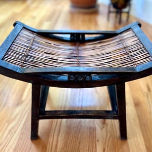 May include: A dark brown wooden stool with a woven bamboo seat. The stool has a curved seat and legs, and is made of dark wood. The bamboo seat is a light brown color. The stool is a decorative piece of furniture.